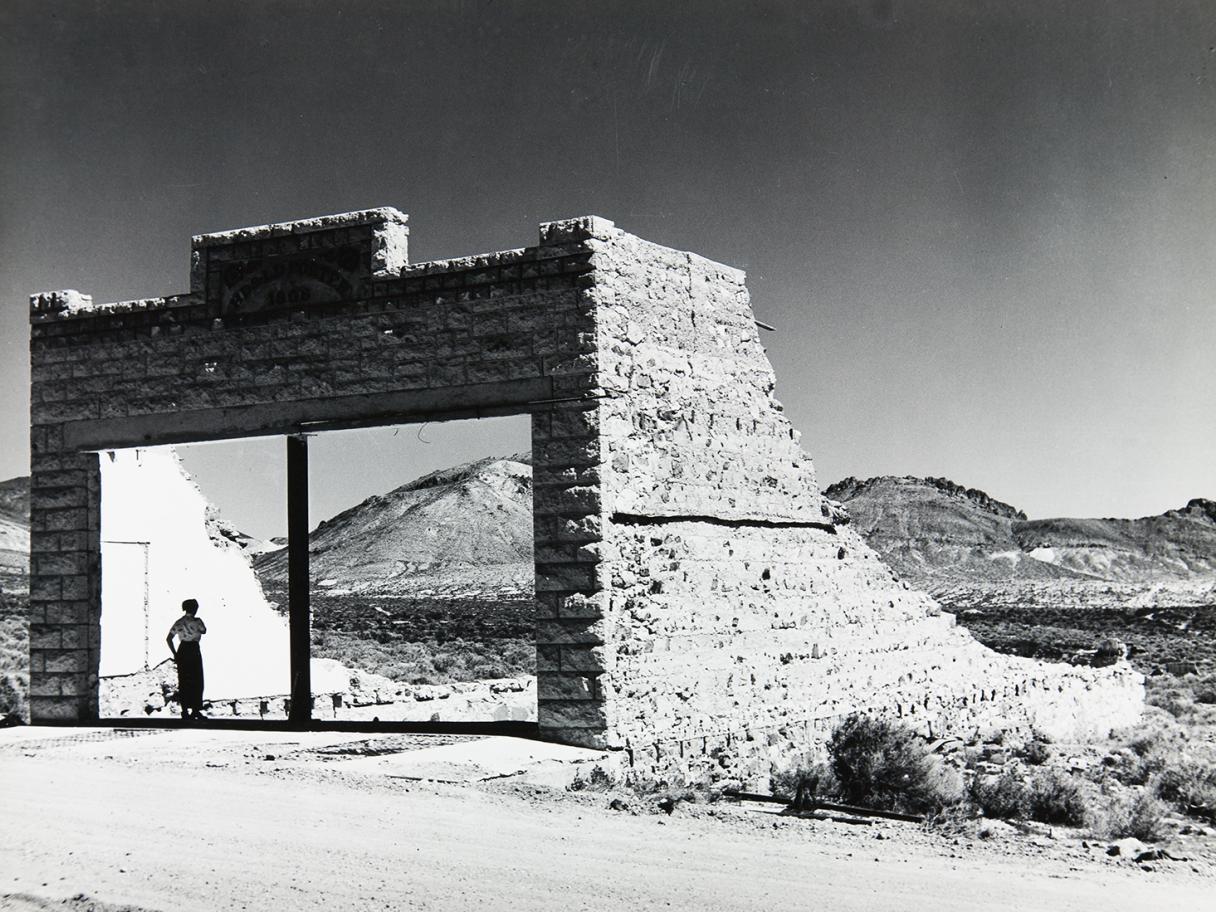 a black and white photograph of a women standing in the large entry to a ruined building made of rocks in a desert