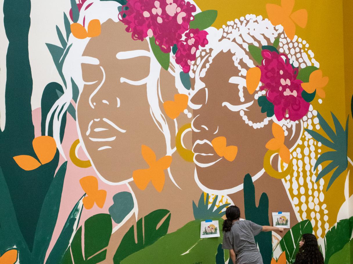 Young women painting a mural of two brown women with pink flowers in their hair.