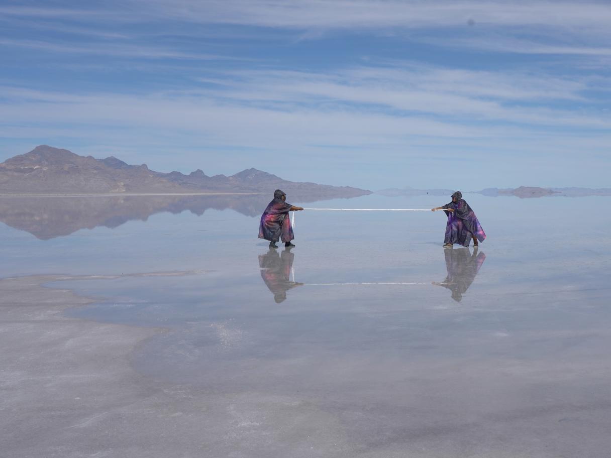Two black cloaked people pulling a string in a reflective lake with a blue sky and shadowy mountain.