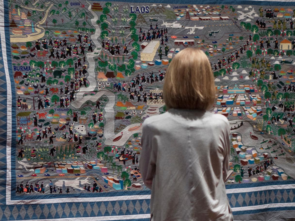 Woman standing in front of a multi colored quilt depicting the migration of the Hmong people, Hmong Story by Bao Lee