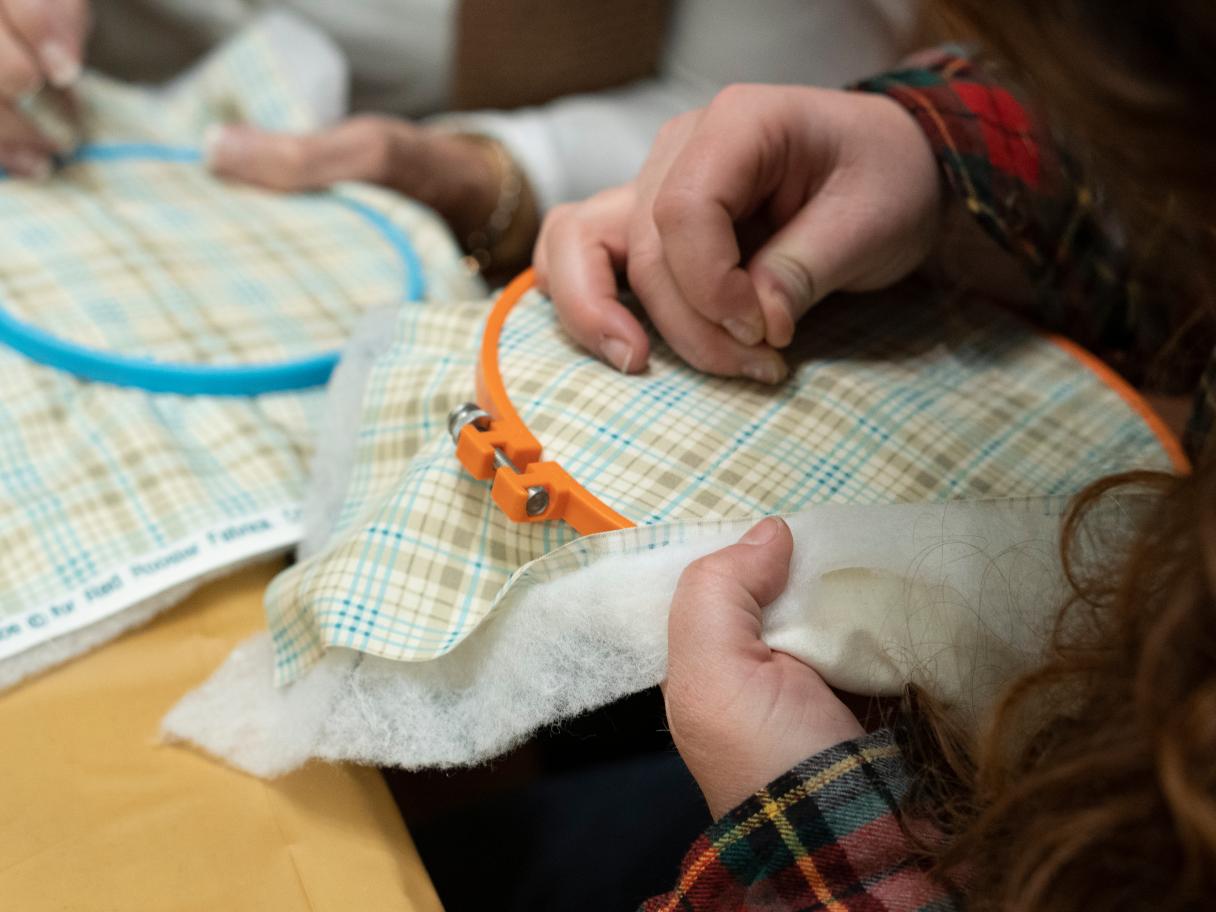 a close up image of a person's hands holding an orange embroidery hoop with plaid fabric in one hand and a needle stitching in the other