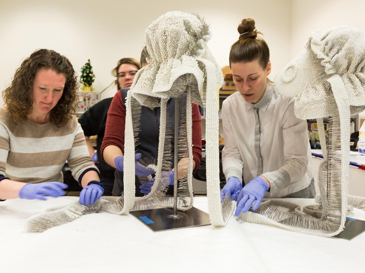 Whitney Tassie and exhibition designer Sarah Palmer work together to prepare two prairie bonnets covered in pearl-headed pins for display