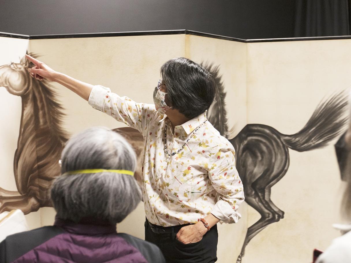 A tall Japanese style screen made of four panels with two large horses, one black one brown painted in large, gestural strokes arcoss the panels. Kimi Hill stands in from on the screen gesturing to a detail for the crowd of people looking on.