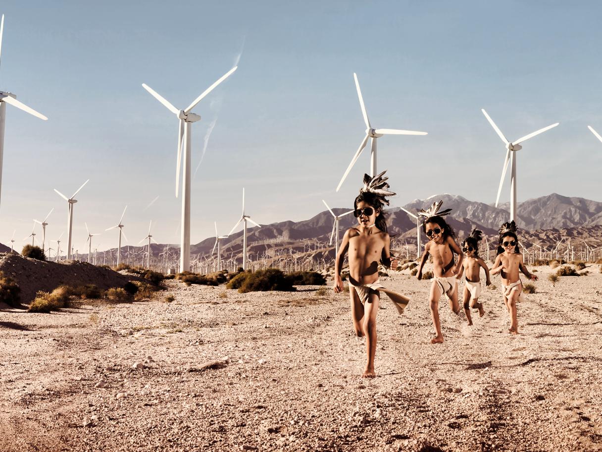 A group of three Indigenous boys in traditional garb running on a desert landscape with wind turbines in the background.