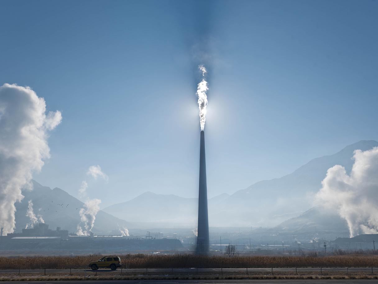 A large smelter with white smoke coming out of it in front of the sun in a blue sky.