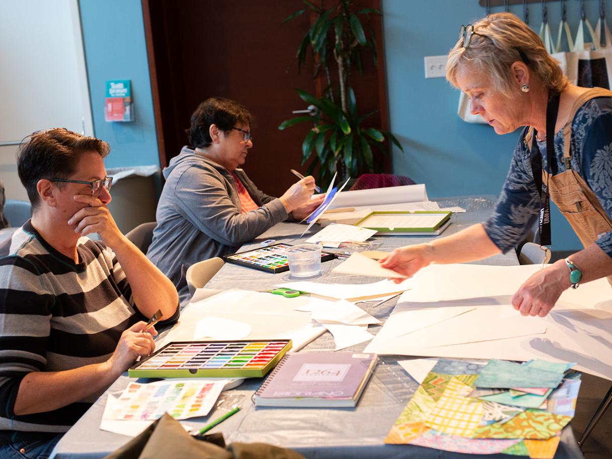 Creative Aging hero Three people site at a long table covered in sheets of paper, there is a large pallet of watercolor paint in front of one of them