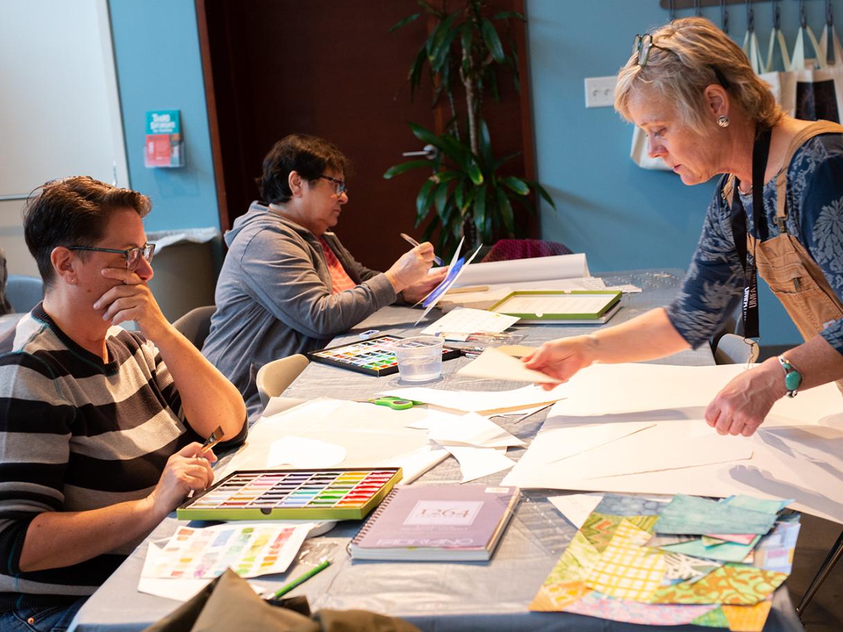 Three people site at a long table covered in sheets of paper, there is a large pallet of watercolor paint in front of one of them