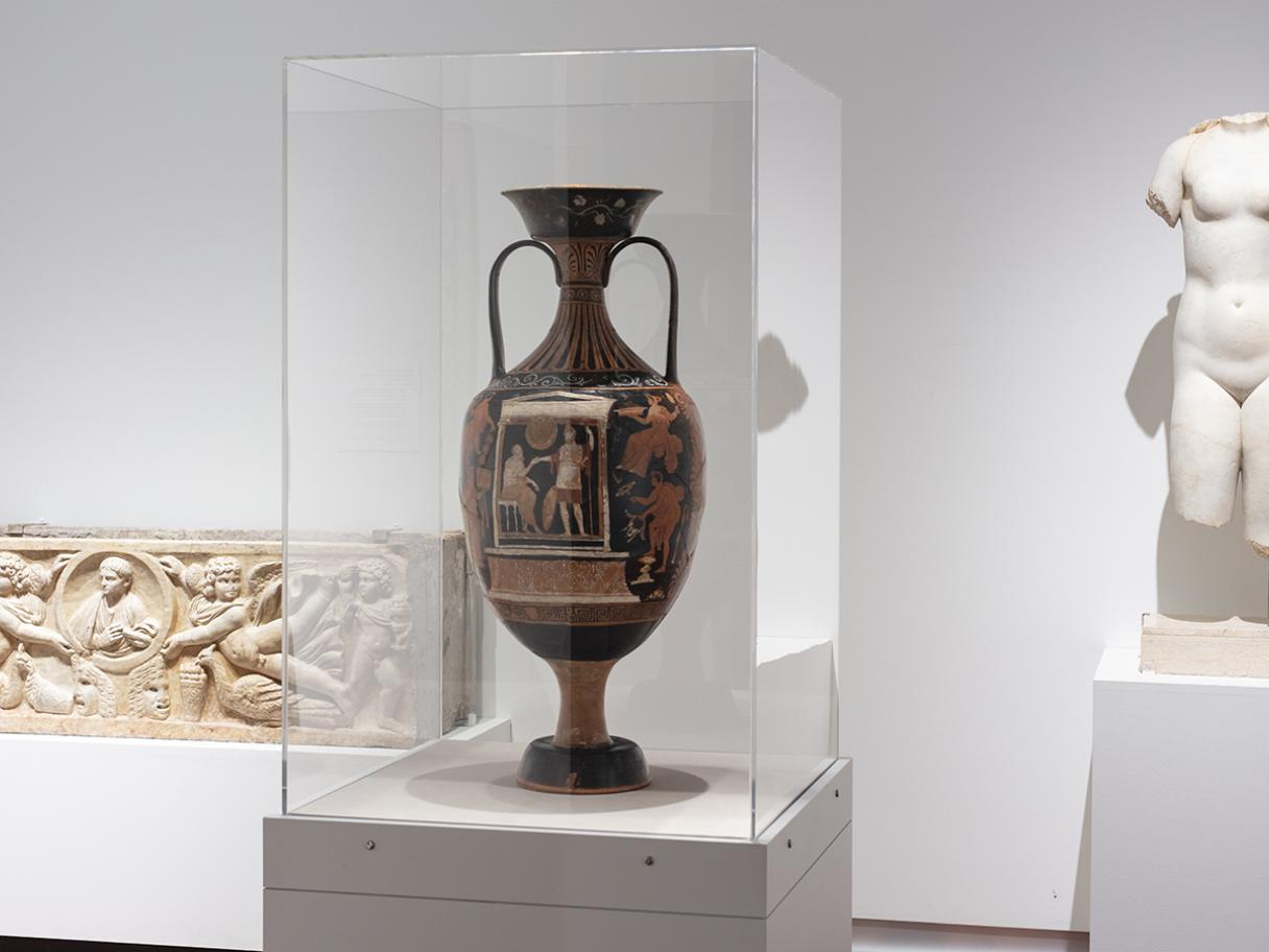 Gallery view of the permanent gallery exhibition "Ancient Mediterranean Art: Res Mortis." In the center of the image is a black amphora with orange drawings, to the left is a ancient greek sarcophagus made of white stone, and to the right is a statue of a Venus with Cupid made of white marble. Venus' head and hands are missing.  
