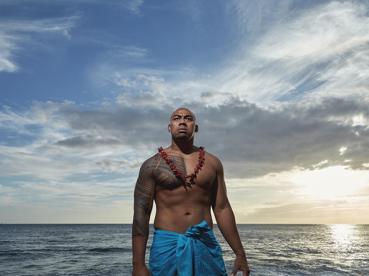A man with a large tattoo covering his shoulder, chest and arm, wearing a blue wrap around his waist, and a red chief necklace stands on a beach looking proud. 