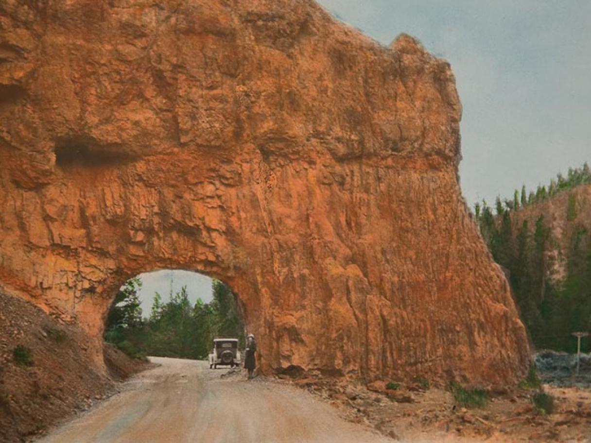 an antique hand colored photograph of a red rock wall wtih a tunnel going through it there is a woman standing next to a car in the tunnel