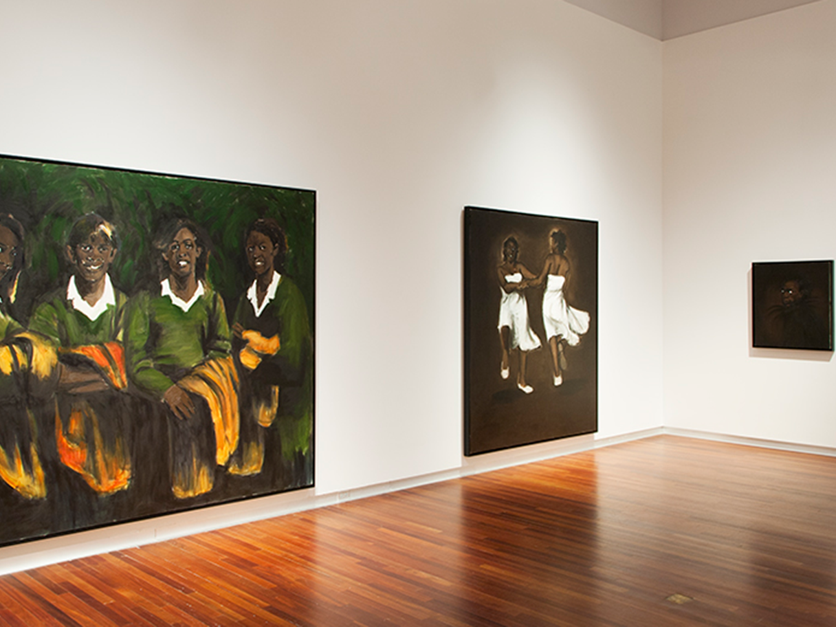 Three dark painting in a gallery with white walls and wood floors. They each show black women smiling or dancing