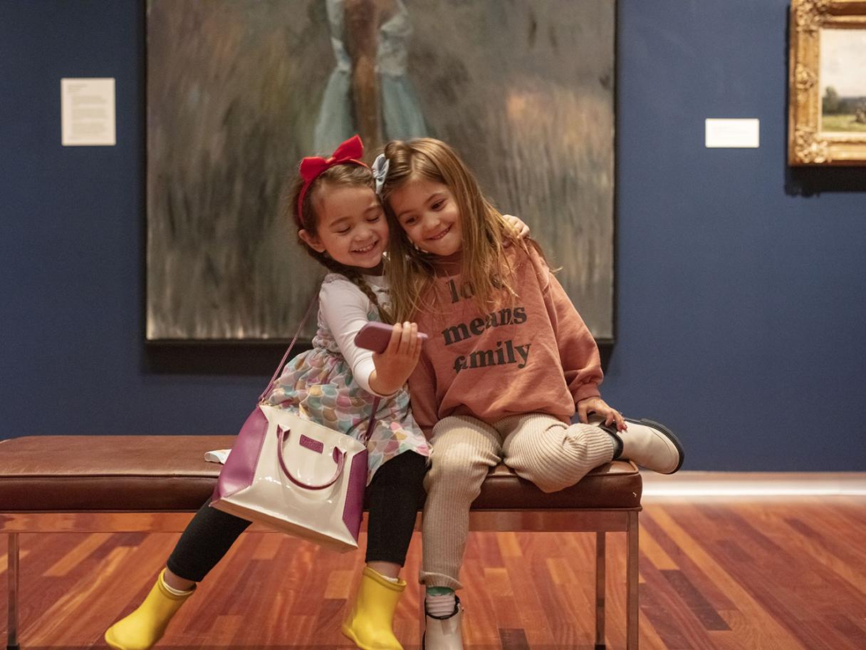 Two little girls sit on a bench in a museum gallery pretending to take a picture with a toy camera