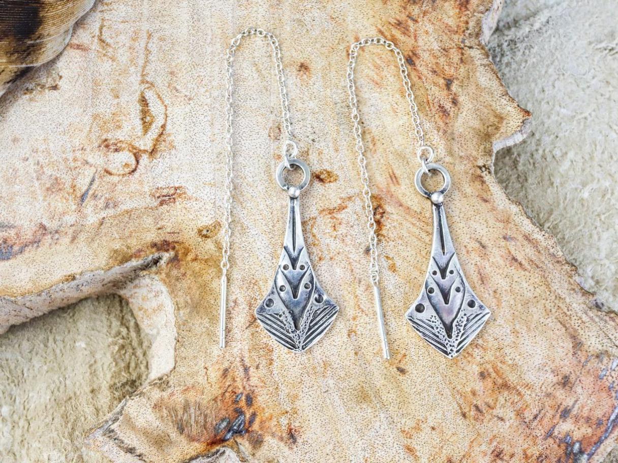 Silver chain earrings with arrow-shaped, stamped pendants laying on a light brown background