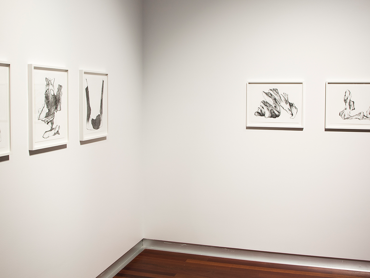 Five black paintings on white backgrounds are mounted on the white walls in a gallery with wooden floors