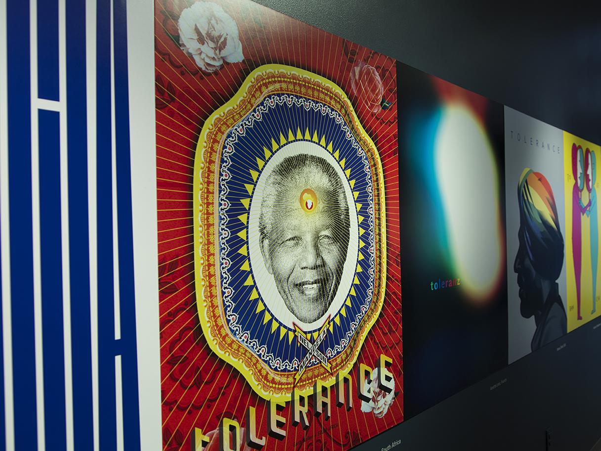 There are multiple posters with different, colorful art lined up next to one another on a black wall. The closest poster says TOLERANCE and has an image Nelson Mandela