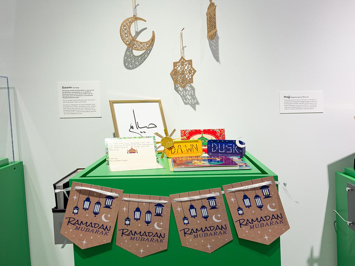 Small items are laid out on a green table. There are small brown flags that say Ramadan lining the front of the table. There are brown wooden cutouts of a crescent moon, star and lantern hanging above the table.