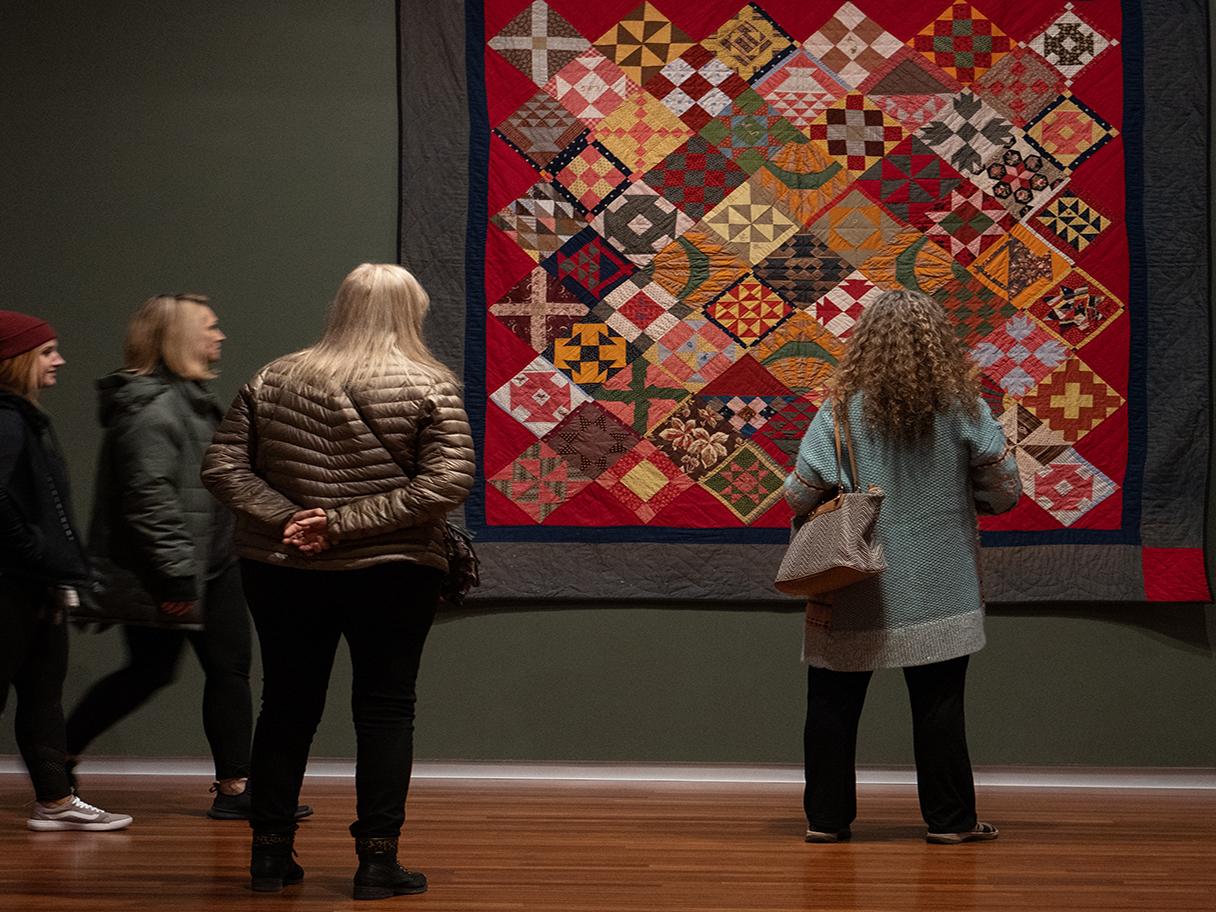 Four women in warm clothes stand and look up at a large, colorful textile on a dark green wall. 