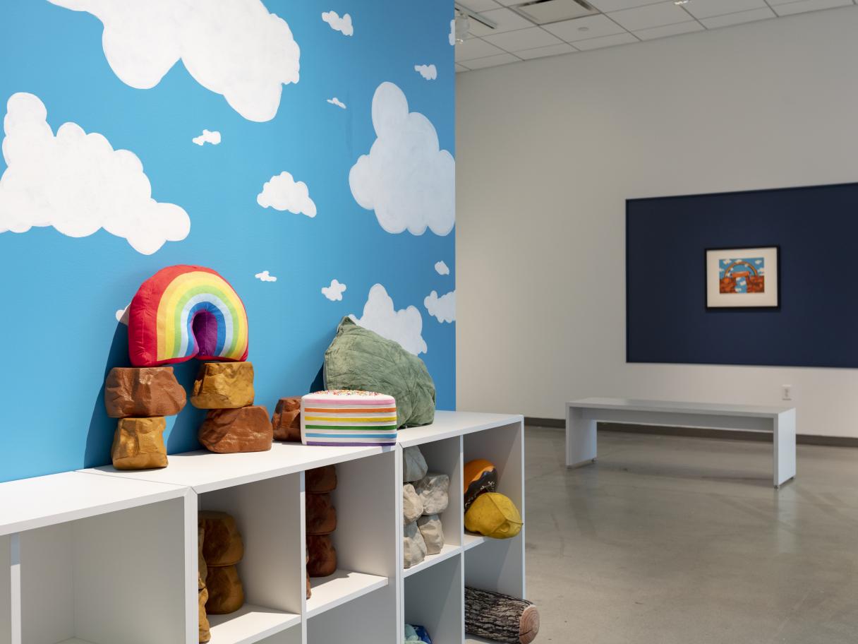 A white shelf with cubbies filled with random, colorful toys. There's a rainbow on top, all against a painted blue sky with white clouds