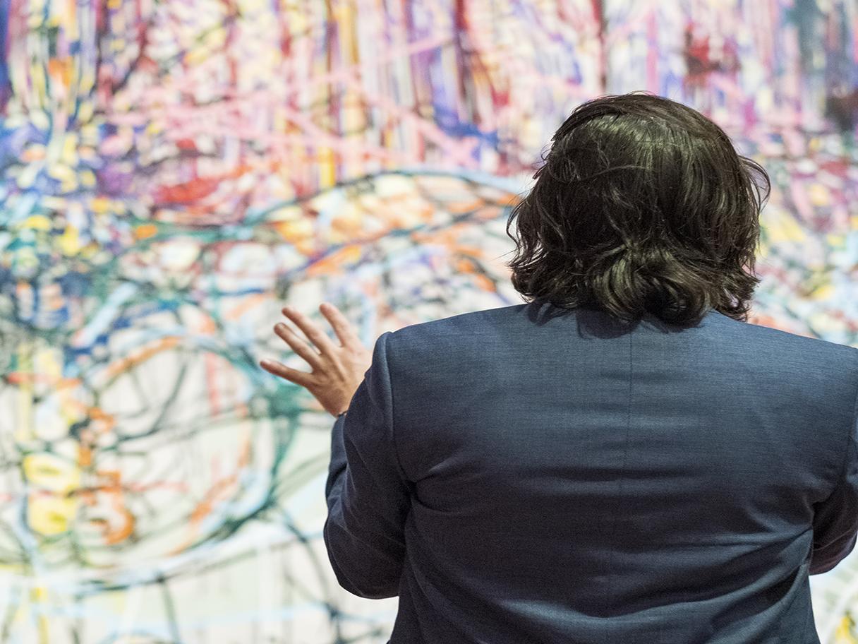 A man in a dark suit stands next to a woman, with their backs to the camera, looking at a colorful piece of art.
