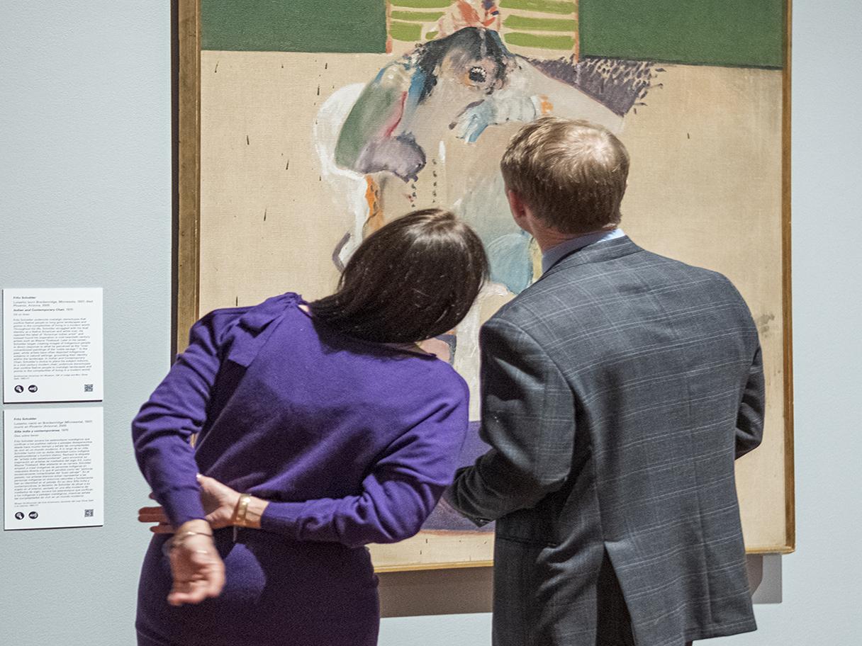 Two people lean toward one another while looking at a painting on the wall. Their backs are to the camera