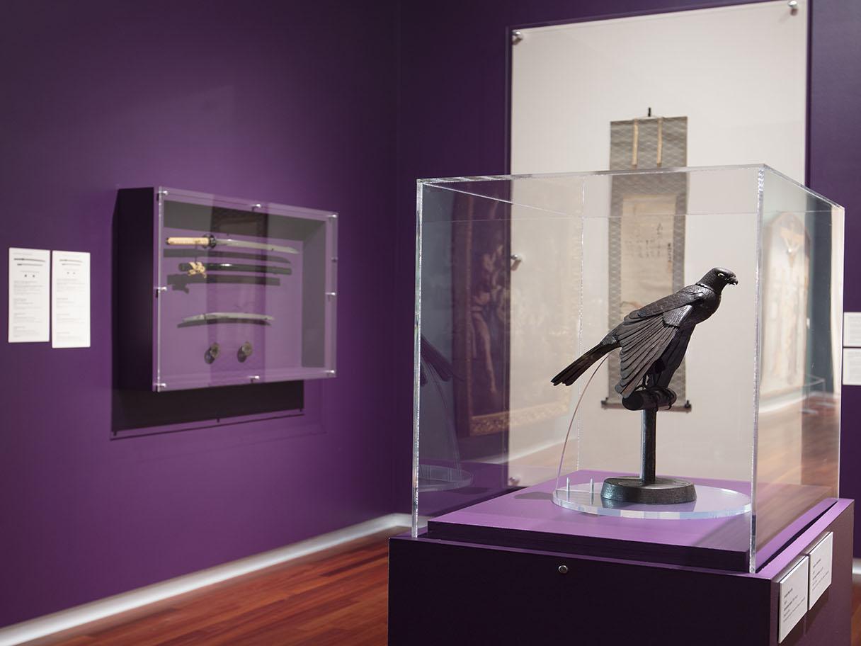 A statue of a sparrow sits in a glass case. There's another glass case in the background, mounted to a purple wall, with swords in it.