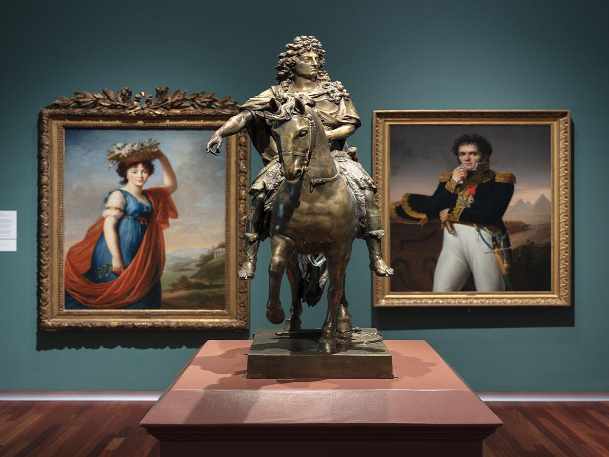 A statue of a man on a horse is on a platform in the foreground. In the background are two portrait paintings in gold frames on a dark green wall