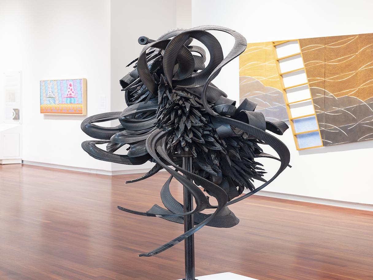 A sculpture made of black tires stands in the middle of a room. There are colorful paintings on the white walls in the background.