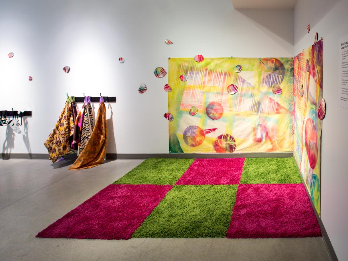 A green and pink checkered, textured rug sits in the corner of a white room. There's a colorful sheet hanging on the joining walls over the rug. Small colorful circles hang from the ceiling. There are colorful smocks hanging on a coat rack on the wall too.