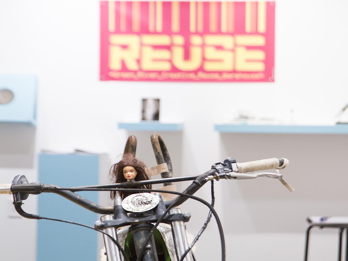 A brunette Barbie with messy hair, sits on an old bike with a black frame and white handlebars. There's a pink sign with cream lettering in the background that says REUSE