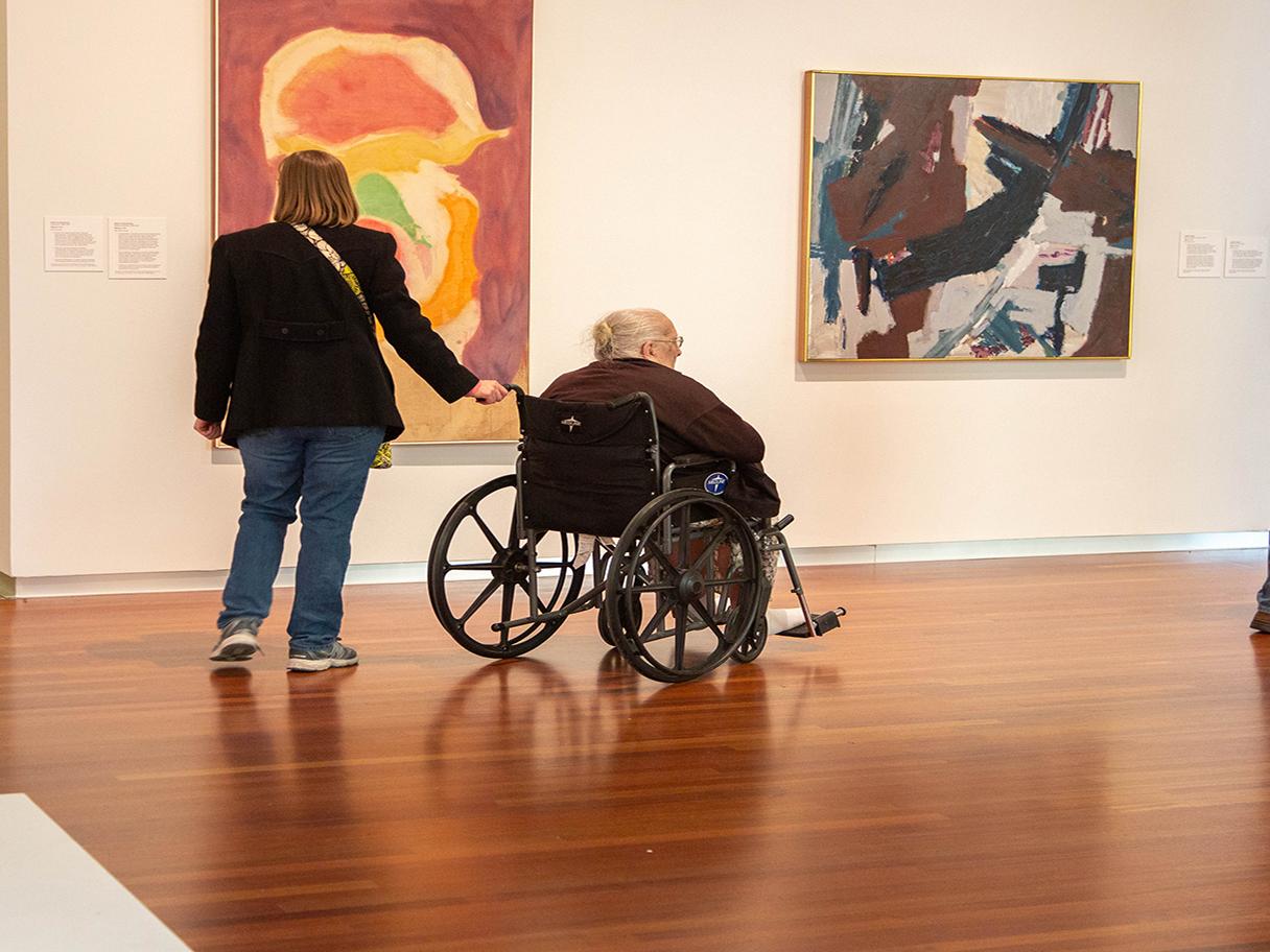 A woman in a dark jacket pushes an older woman in a wheel chair. They are looking at colorful paintings on a white wall. 