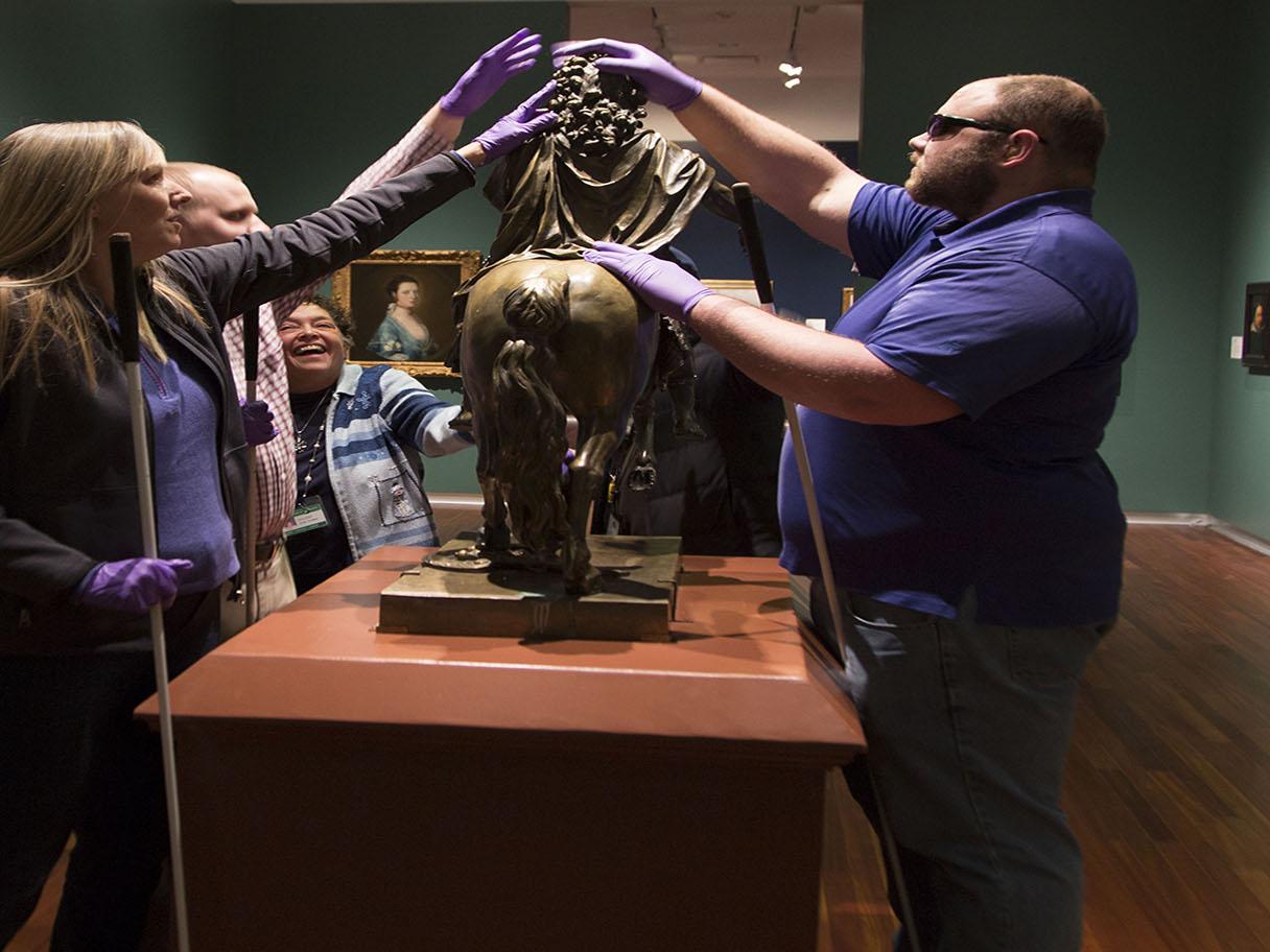 A group of people with purple gloves on touch a small, bronze statue of a horse 
