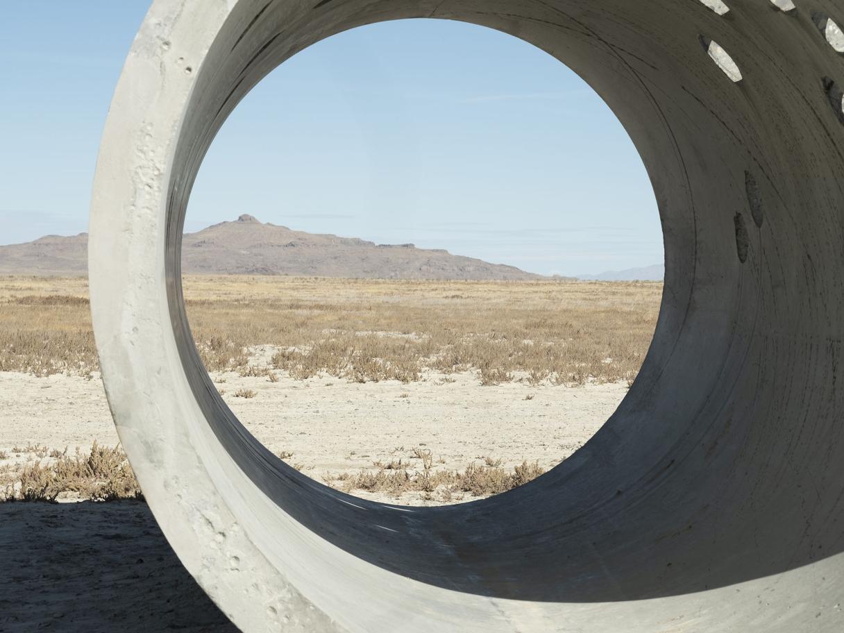 A large concrete cylinder sits in the middle of an open desert landscape. There is tan sand and dry, green plants on the ground. A mountain sits in the background against a light blue sky.