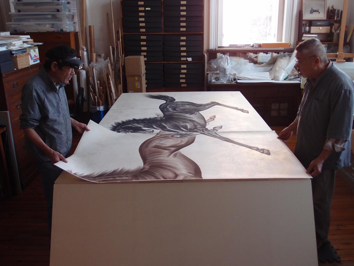 A photo of two men standing on either side of a large table, touching paper with paintings of two horses.