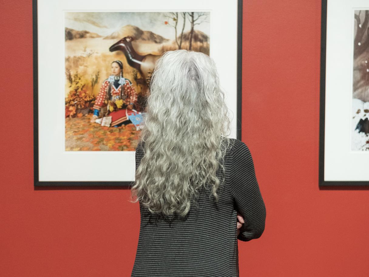 A photo of a woman with long light grey hair staring at a framed painting of a woman and camel on a bright red wall.