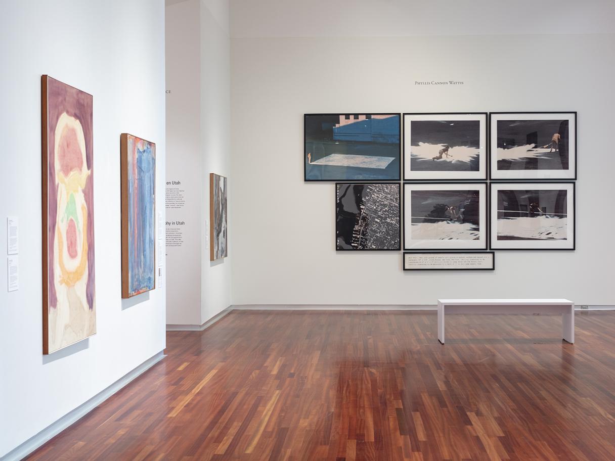 A photo of a large gallery room with white walls and wood floors. There are three painted canvases hanging on the walls to the left. There are several, large framed images on the far wall with a white bench placed in the middle of the room in front of them.