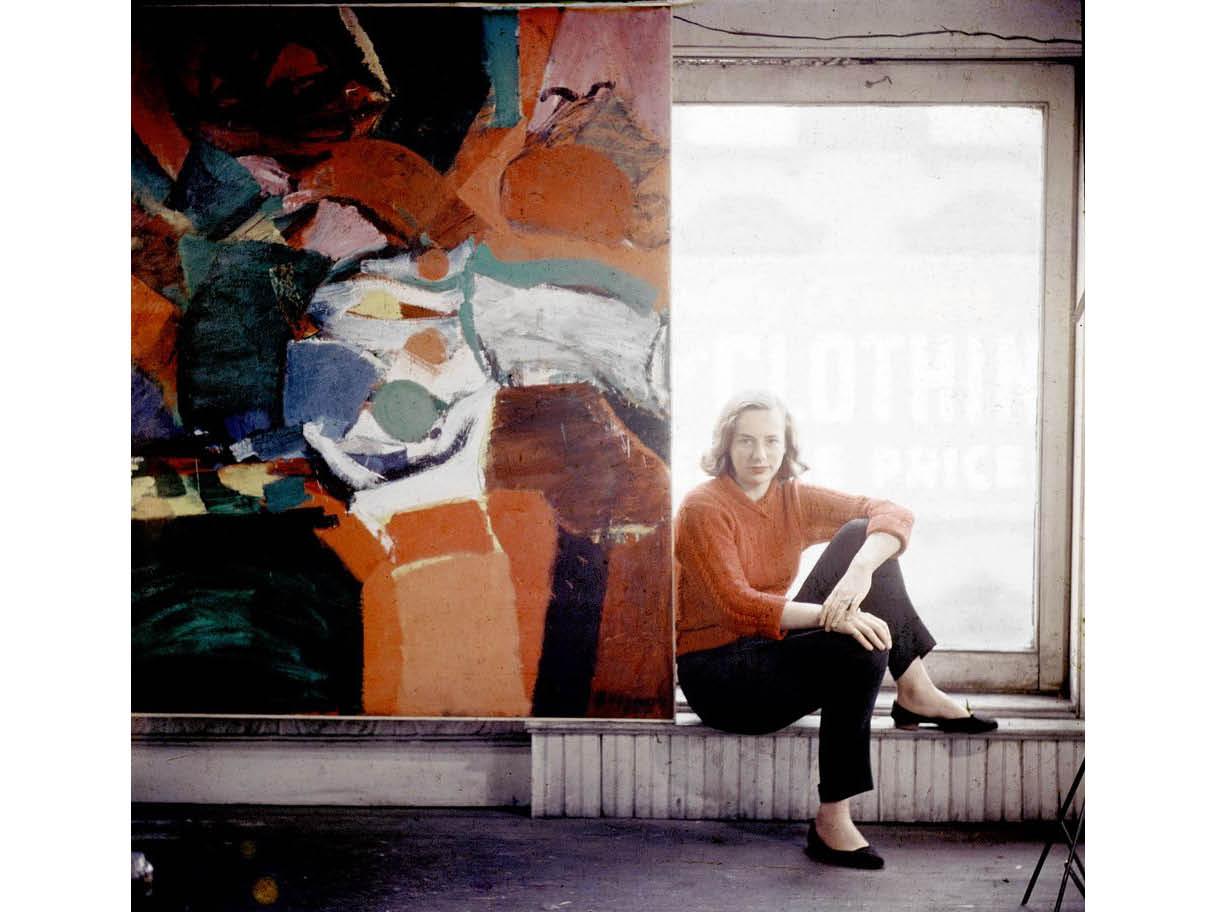A photo of a woman in black pants and a red shirt, sitting in front of a window next to a large painted canvas.