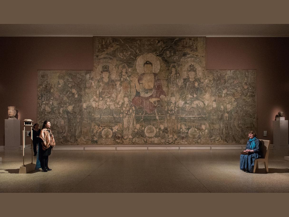 A photograph of two people on opposites sides of a large room; one is sitting in a blue dress, and the other is standing and singing. There's a large mural with eastern deities in the background.
