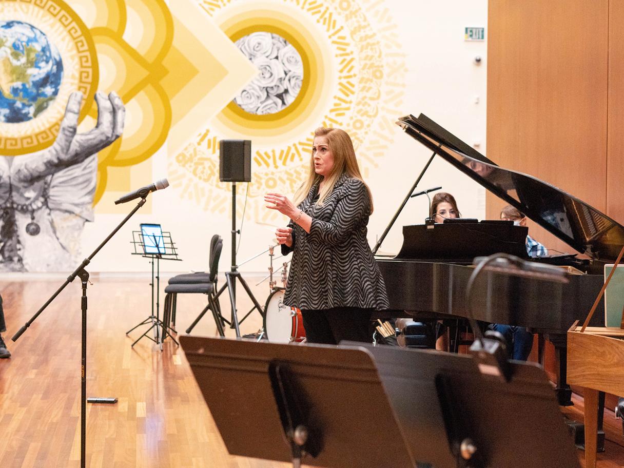 A white woman with long hair wears a dark shirt and pants. She's singing into a microphone in front of a gold mural. There is a piano and a woman playing the piano behind her.
