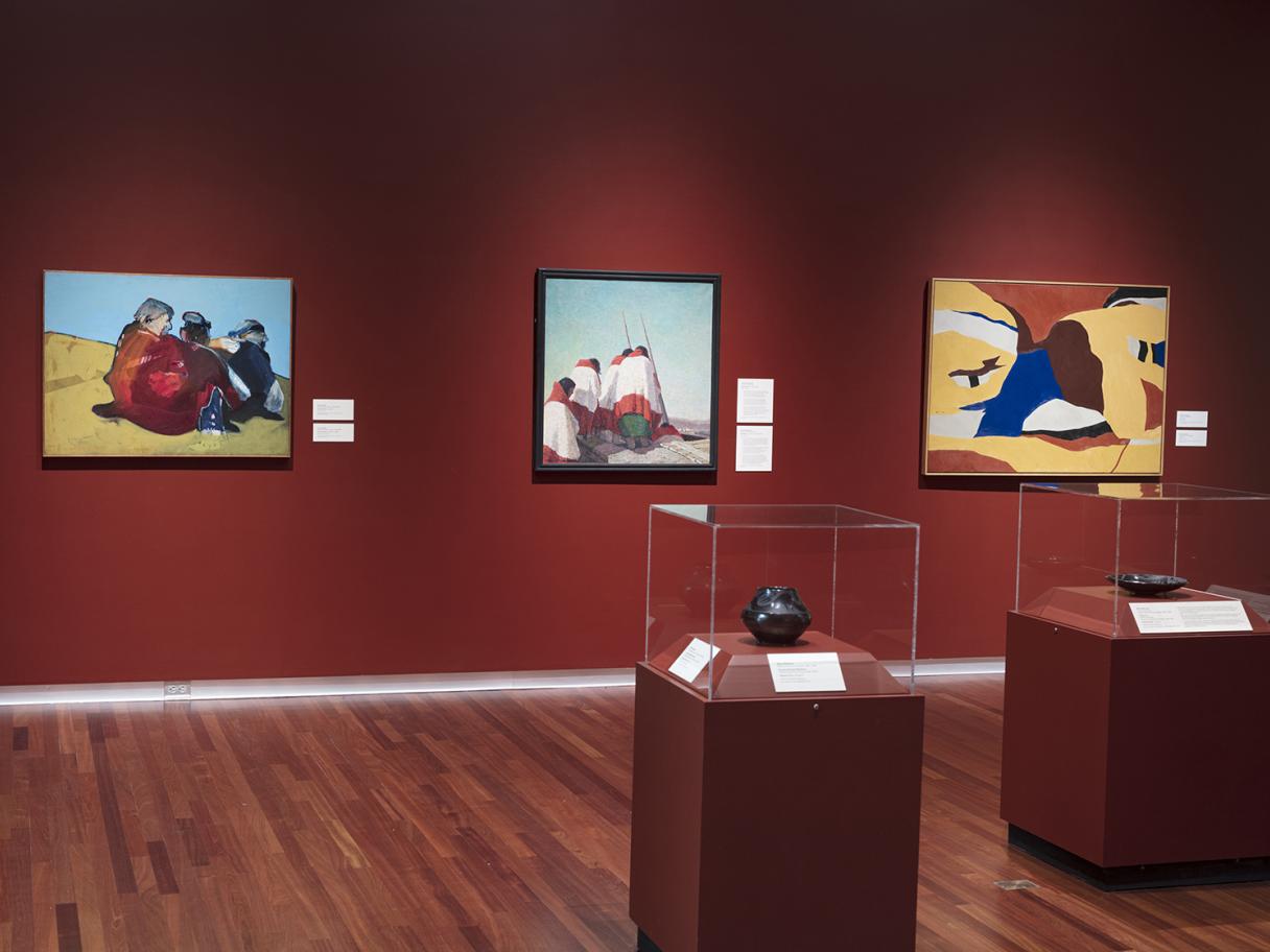 Three painted canvases line the back, burgundy-colored wall. There are two dark pieces of pottery in glass display cases on similar-colored stands in the middle of the room. 