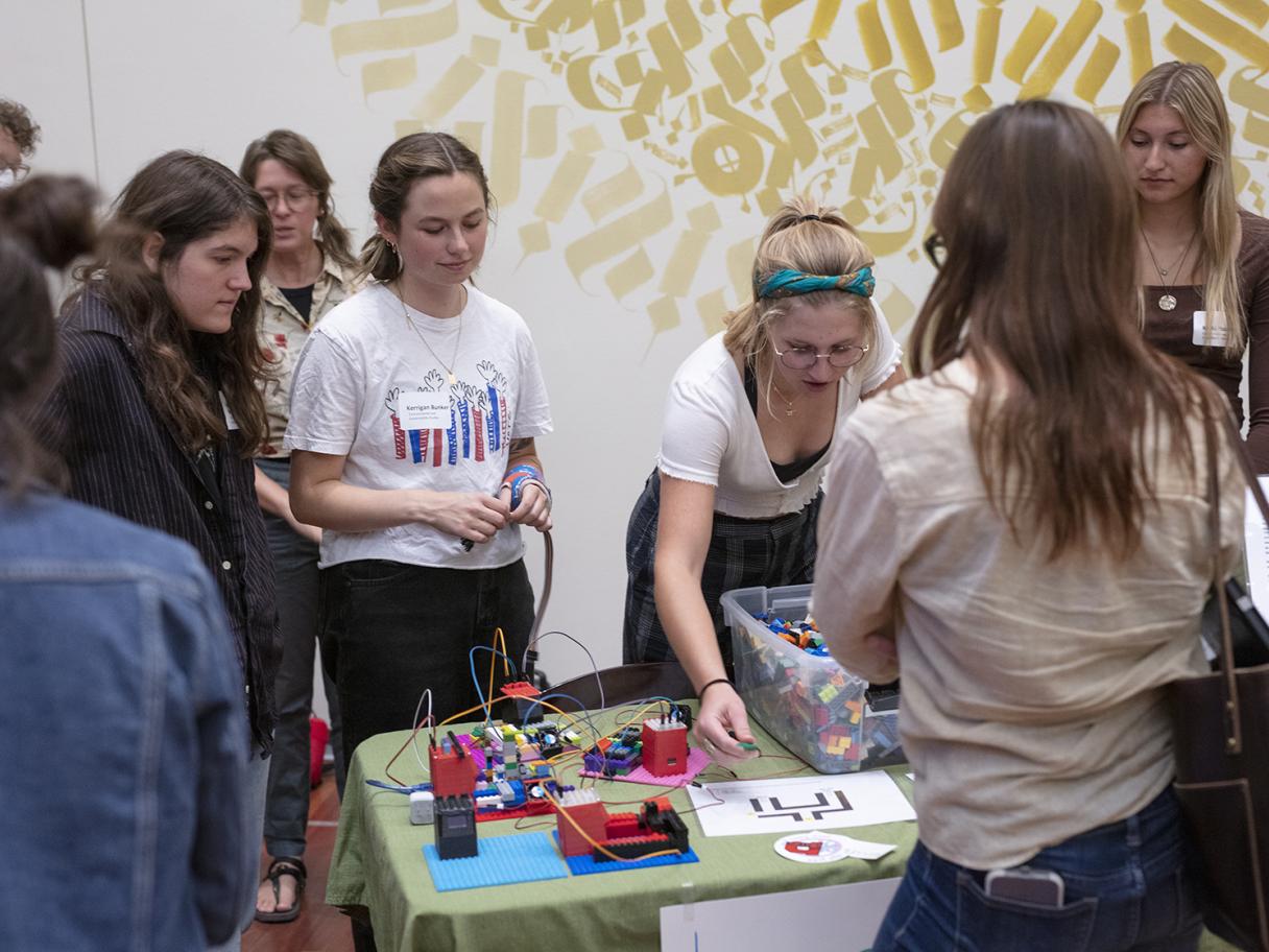 A group of women stand around a table with clips and wires. One woman is touching the objects on the table as she looks down. The rest watch over her. There is part of a gold patterned mural painted on the wall behind them.