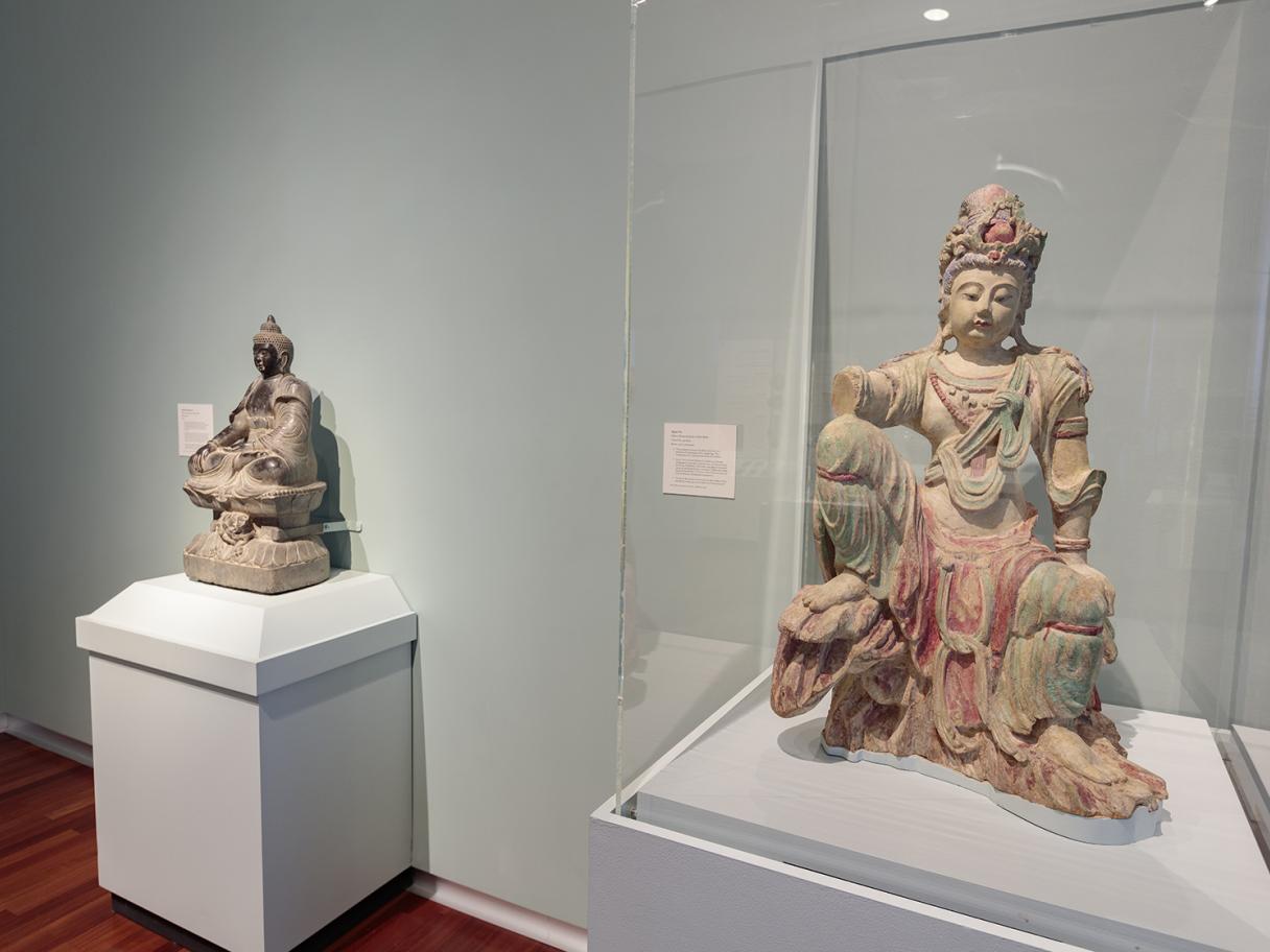 Two carved deities sit on separate mint-colored stands. One is in a glass display case.