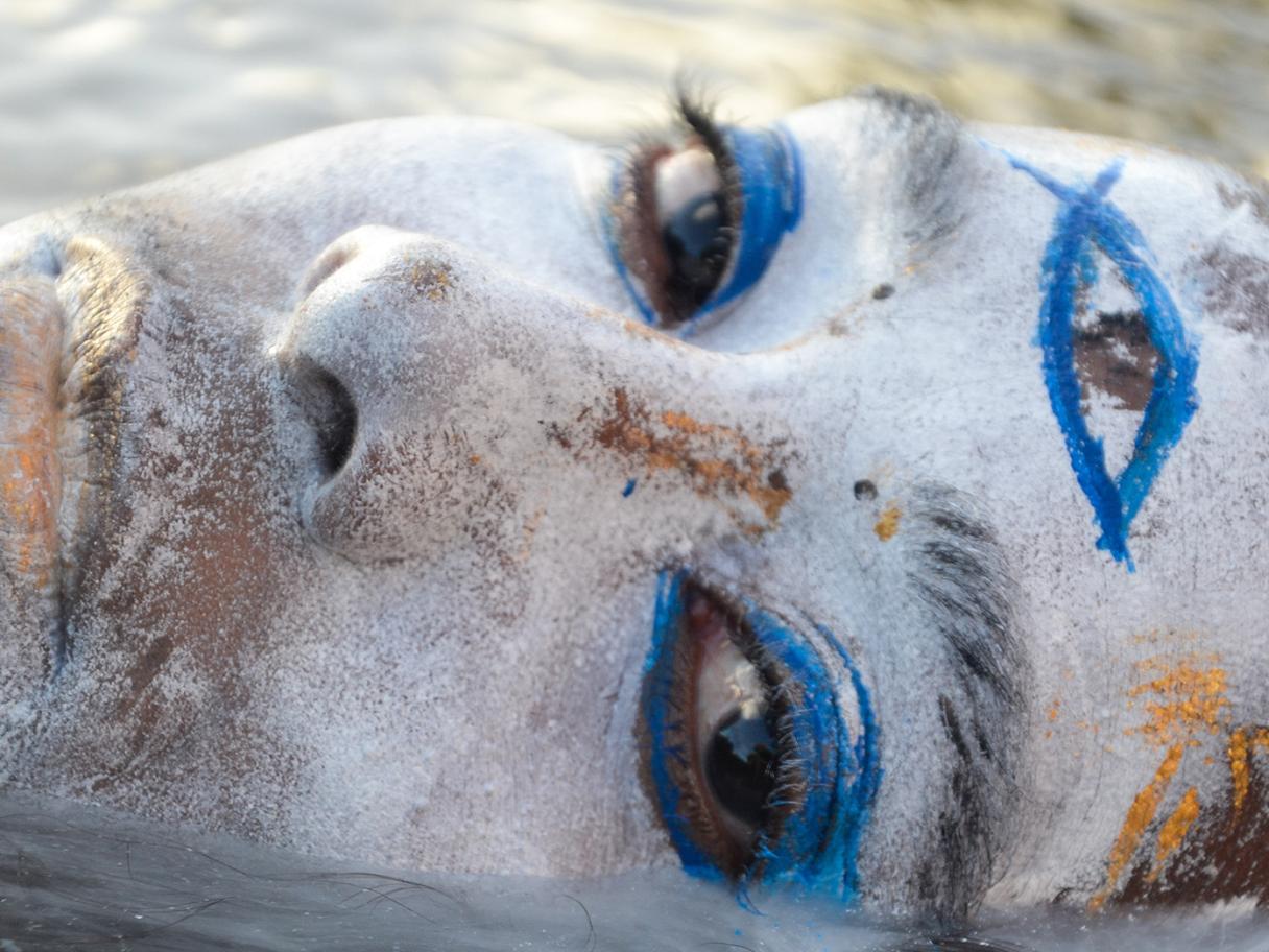 A close up image of a woman's head that's painted white with blue on the eyes and an eye drawn on her forhead, laying in the water and looking at the camera.