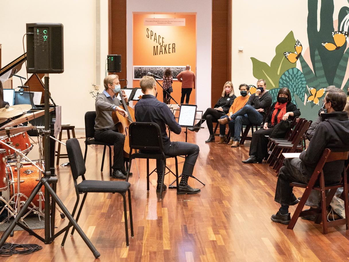 Two white men sit on chairs, wearing masks and playing string instruments in front of a seated audience of several different people.