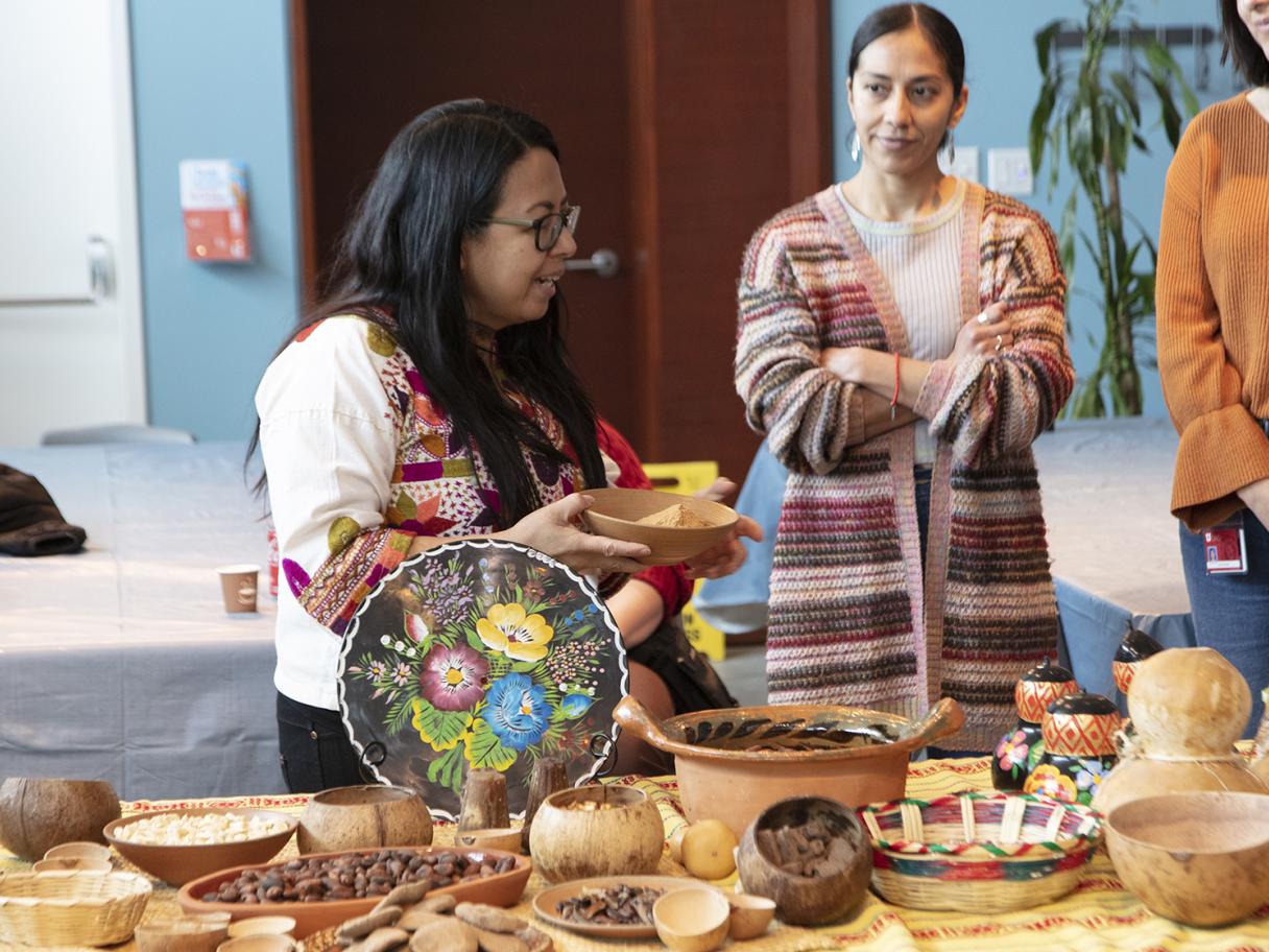 A woman holds a bowl and talks to people standing and sitting around her. There's a table in front of the with different shape, color and size bowls with cacao beans in them.