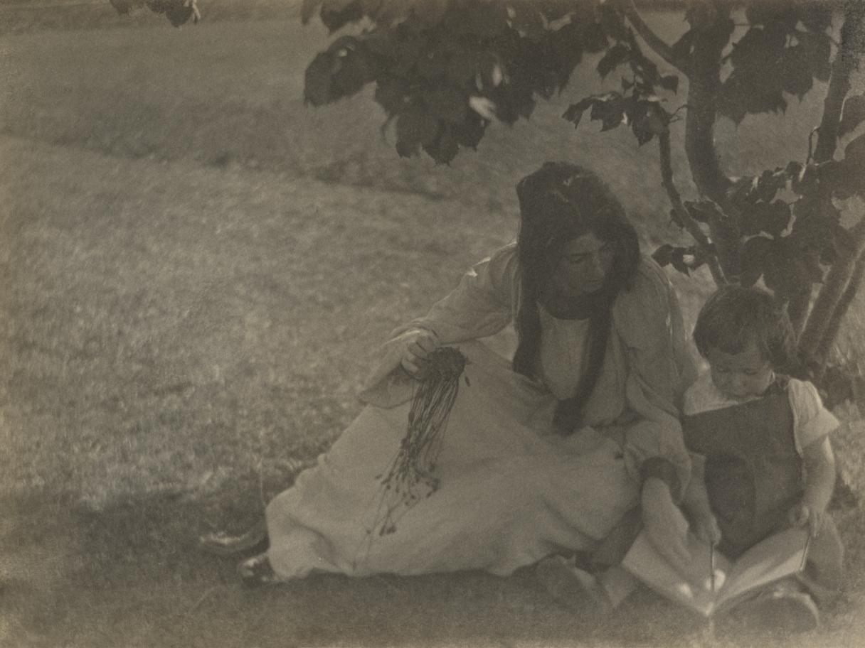 A black and white photo of a woman in a white dress sitting on the ground beneath a tree with a small child looking at a book.