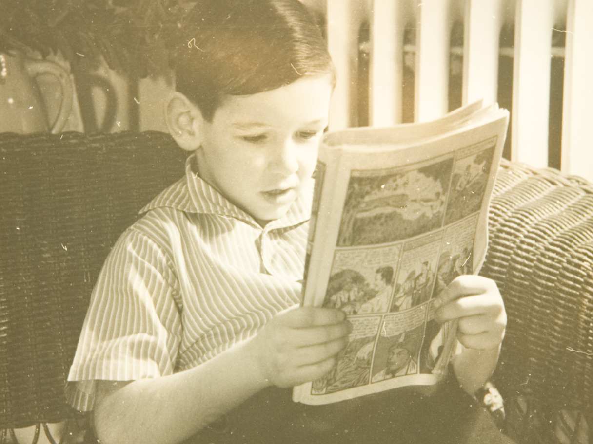 A black and white photo of a young boy reading a comic book. 