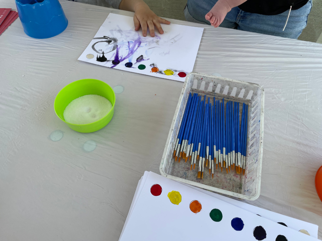 A table with paint brushes and white paper with watercolor dots on them. At the top of the photograph a child's hand smudges the paint. 