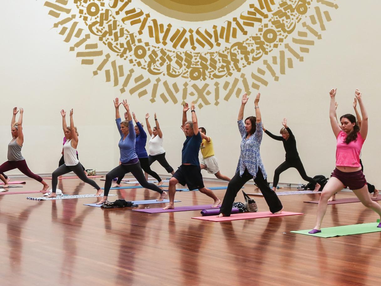A group of people practice yoga in the UMFA Great Hall. A gold mural is in the background.