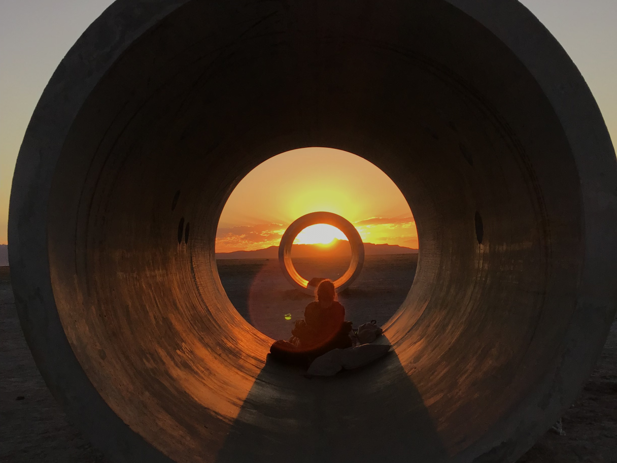 Two concrete tunnels line up with the setting sun. A person sits silhouetted between the two tunnels surrounded by warm orange light.