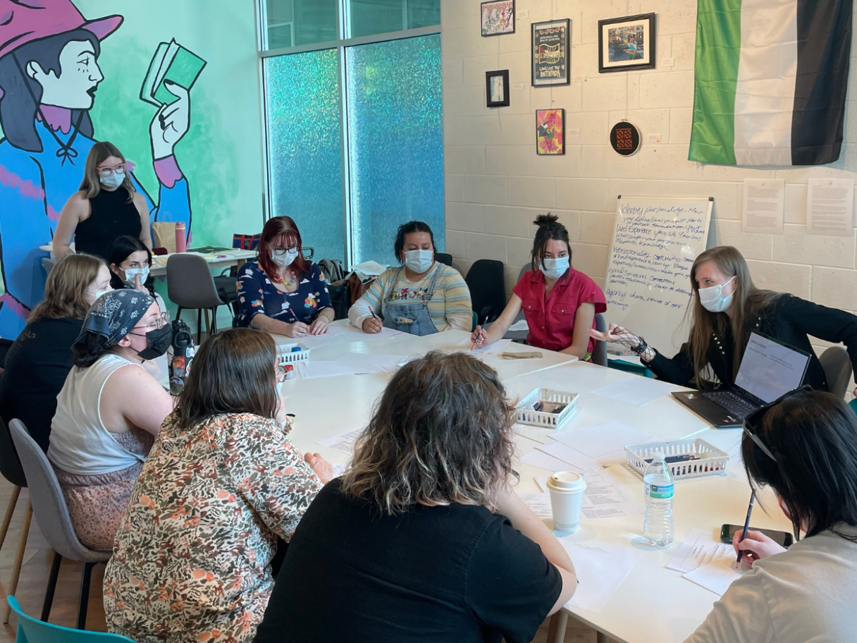 A group of people in masks sit around a table. A mural of a woman reading a book is in the background.